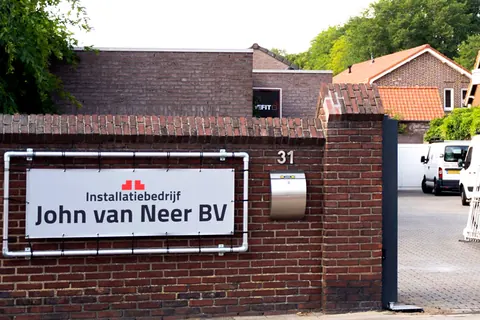 Poort van Installatiebedrijf John van Neer BV met het bedrijfsbord op de muur en bedrijfsbussen op het terrein